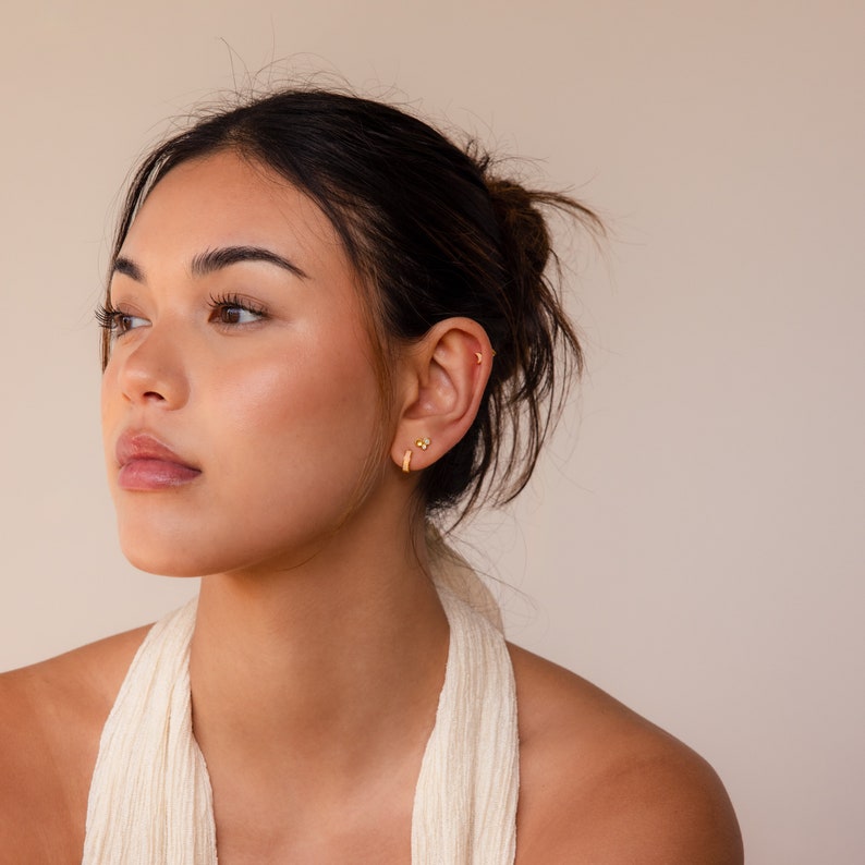 May include: A woman with long dark hair wearing a white halter top and gold hoop earrings. She is looking to the side with a neutral expression.