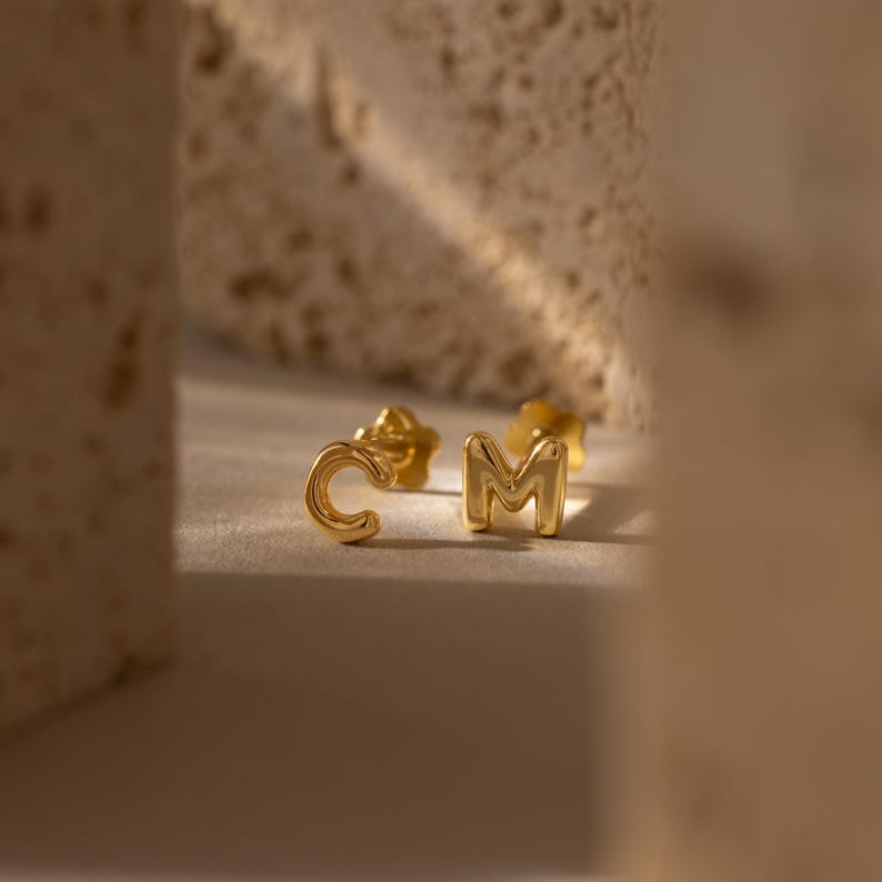 May include: A pair of gold-coloured stud earrings, one shaped like the letter "C" and the other like the letter "M". The earrings are small and have a shiny finish. The background is a neutral colour, with soft lighting.