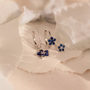 May include: A pair of silver hoop earrings with a dangling flower charm. The flower charm is made of blue gemstones and silver.