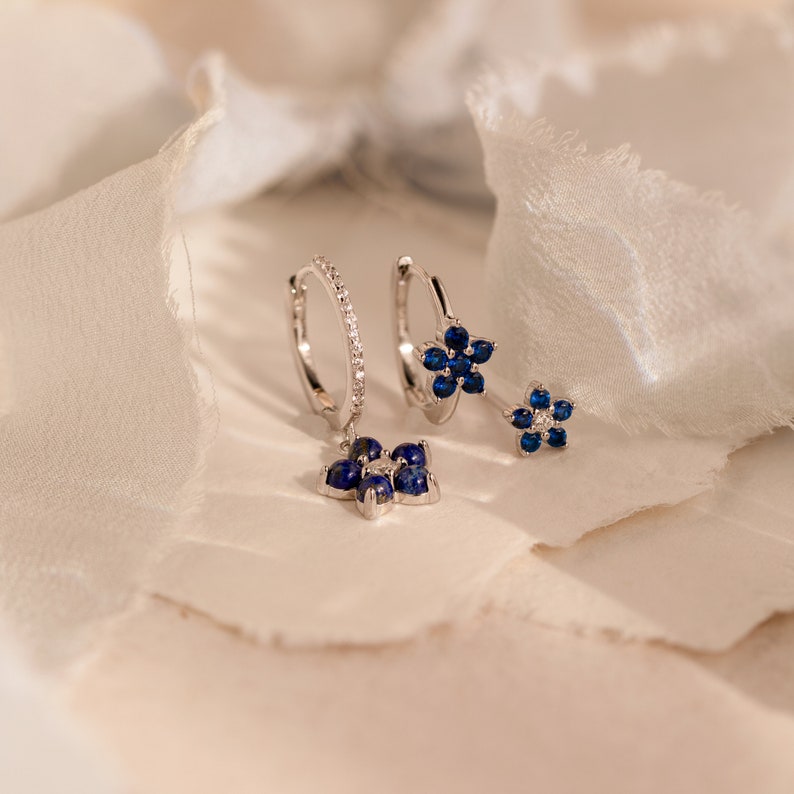 May include: A pair of silver hoop earrings with a dangling flower charm. The flower charm is made of blue gemstones and silver.