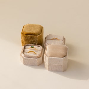 May include: Three velvet ring boxes, one brown and two beige, with rings inside. The boxes are square and have a raised edge around the top.