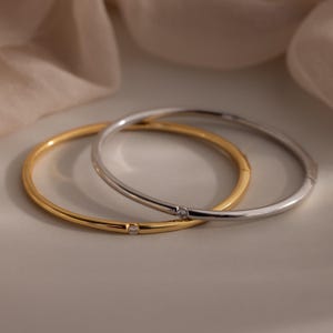 Puede incluir: Dos elegantes brazaletes, uno dorado y otro plateado, cada uno con una pequeña y brillante gema. Los brazaletes se exhiben sobre una superficie neutra, con una tela suave y drapeada en el fondo, creando una estética sofisticada y minimalista.