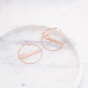 May include: A pair of gold hoop earrings with the name "Lauren" written in cursive on each hoop.