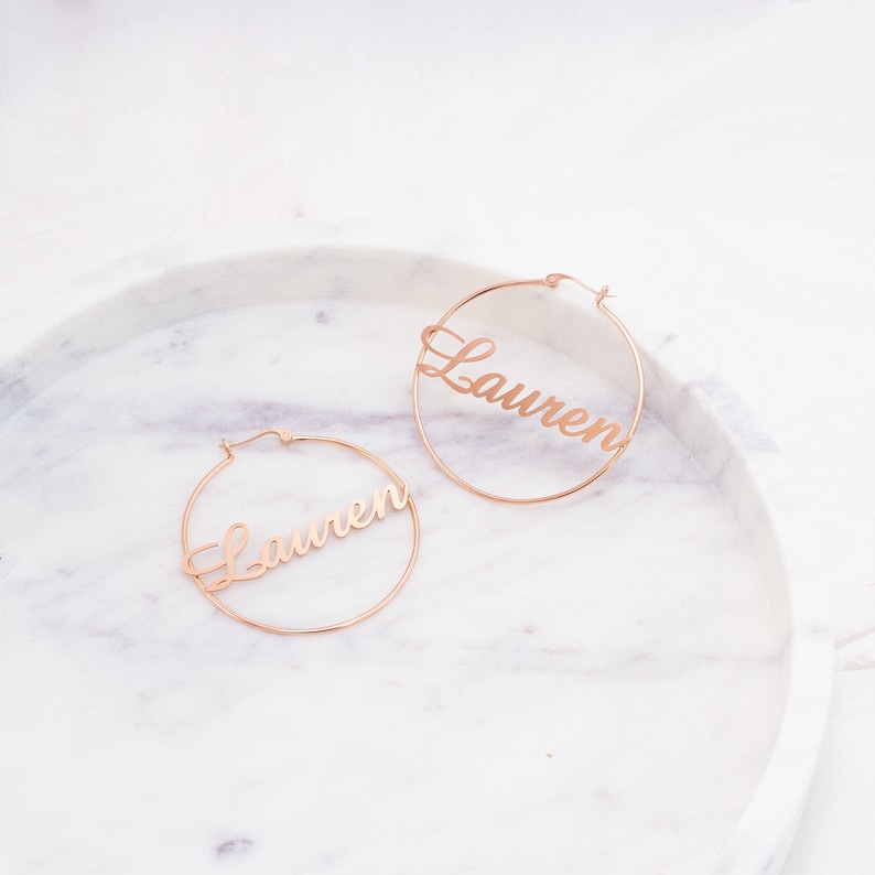 May include: A pair of gold hoop earrings with the name "Lauren" written in cursive on each hoop.