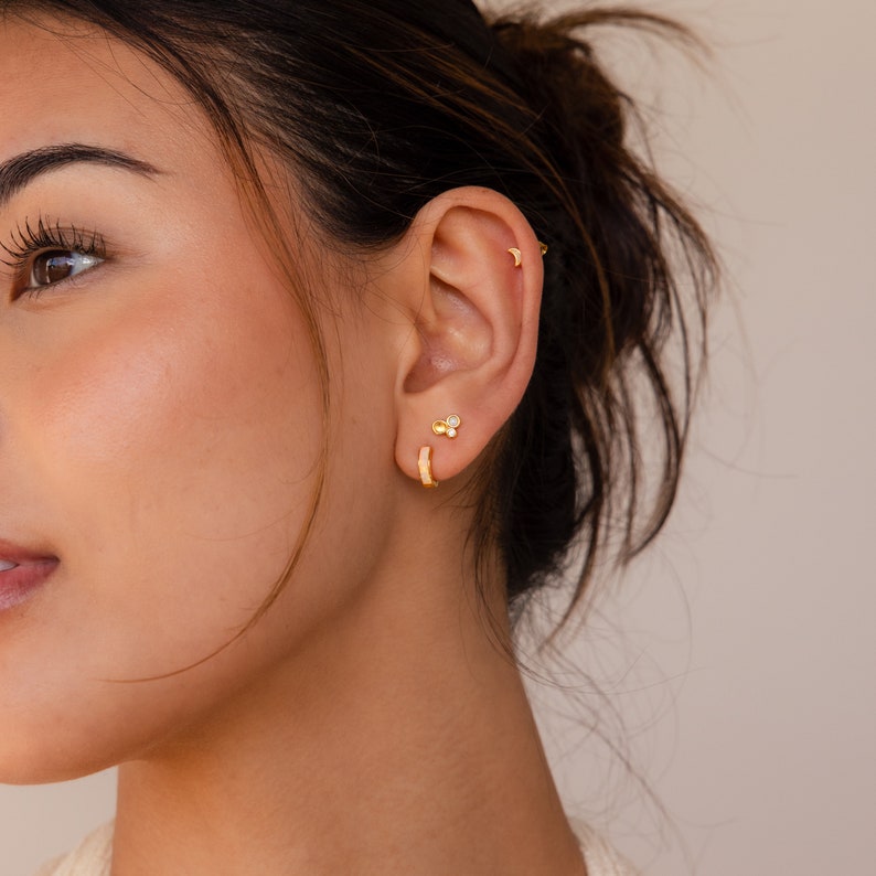 May include: A woman wearing gold hoop earrings and a small gold stud earring with three small stones. The earrings are in her left ear.