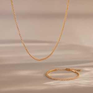 May include: A gold-colored necklace and bracelet set. The jewelry features a delicate design with small, evenly spaced embellishments. The necklace hangs in a gentle curve, while the bracelet is positioned below, both against a soft, neutral background.
