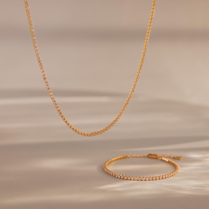 May include: A gold-colored necklace and bracelet set. The jewelry features a delicate design with small, evenly spaced embellishments. The necklace hangs in a gentle curve, while the bracelet is positioned below, both against a soft, neutral background.