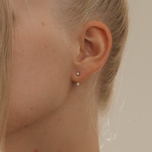 Puede incluir: Primer plano de una oreja con dos pendientes pequeños. Los pendientes son de color dorado con una pequeña gema blanca cuadrada. Los pendientes son de diseño sencillo y minimalista.