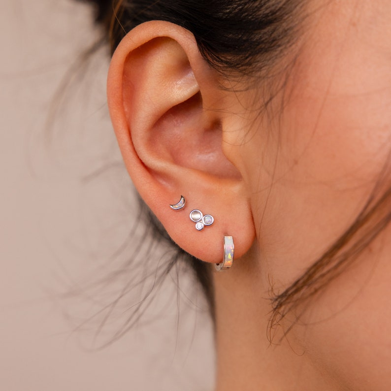 May include: A person's ear with three silver earrings. The earrings are a crescent moon, a cluster of three small round stones, and a hoop earring.