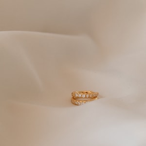 May include: Two gold-colored hoop earrings, each encrusted with small, clear gemstones. The earrings are positioned on a cream-colored, slightly textured surface, creating a minimalist aesthetic. The gemstones sparkle, reflecting light.
