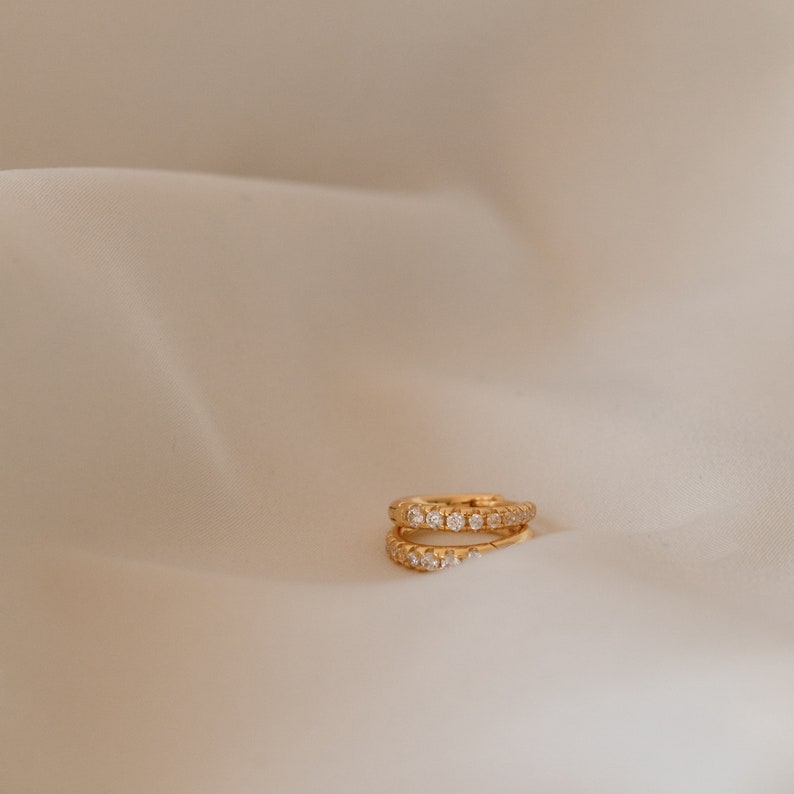 May include: Two gold-colored hoop earrings, each encrusted with small, clear gemstones. The earrings are positioned on a cream-colored, slightly textured surface, creating a minimalist aesthetic. The gemstones sparkle, reflecting light.