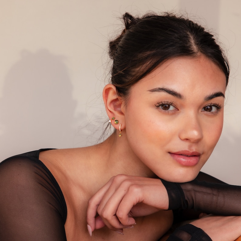 May include: A woman wearing gold hoop earrings with a small green gemstone and a gold dangle earring with a small green gemstone. She is wearing a black mesh top.