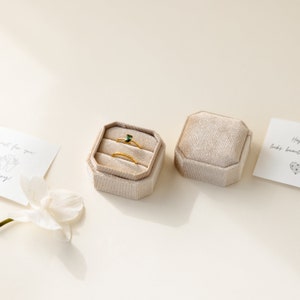 May include: Two gold rings and a green gemstone ring are displayed in a beige velvet ring box. The box is open and the rings are visible. The box is on a white surface.