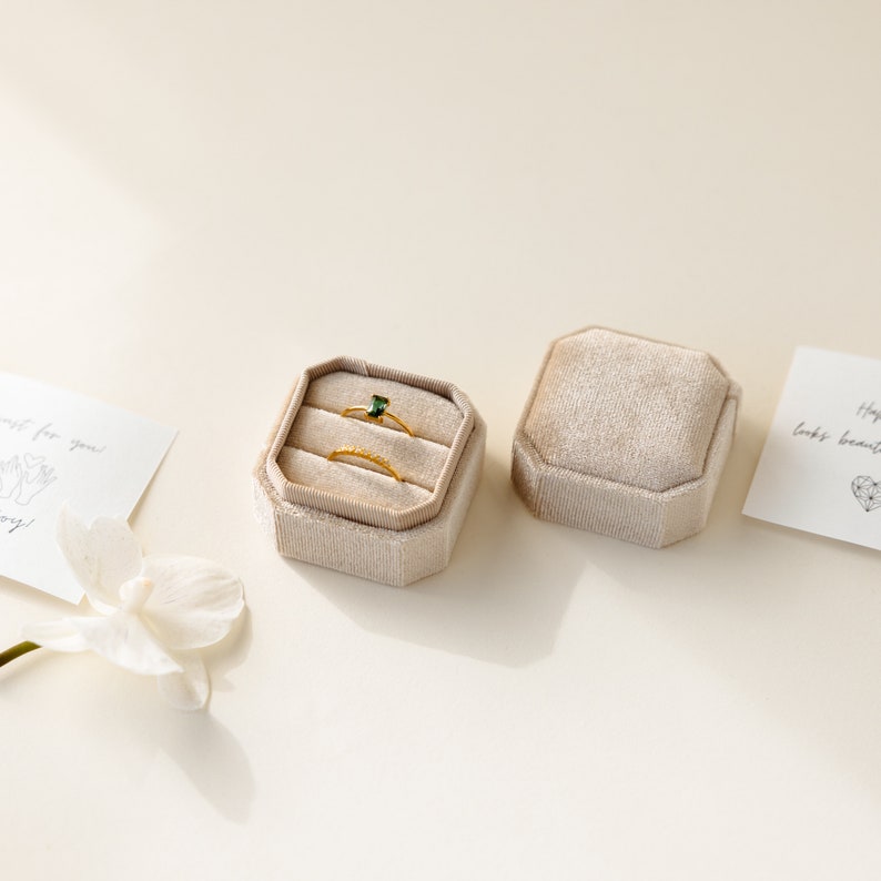 May include: Two gold rings and a green gemstone ring are displayed in a beige velvet ring box. The box is open and the rings are visible. The box is on a white surface.