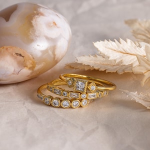 May include: Three gold rings with clear gemstones. The rings are stacked on top of each other, showcasing different designs. One ring features a square gemstone, while others have round and rectangular stones. The background includes a decorative stone and dried leaves.