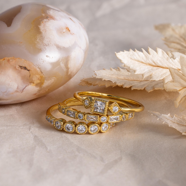 May include: Three gold rings with clear gemstones. The rings are stacked on top of each other, showcasing different designs. One ring features a square gemstone, while others have round and rectangular stones. The background includes a decorative stone and dried leaves.