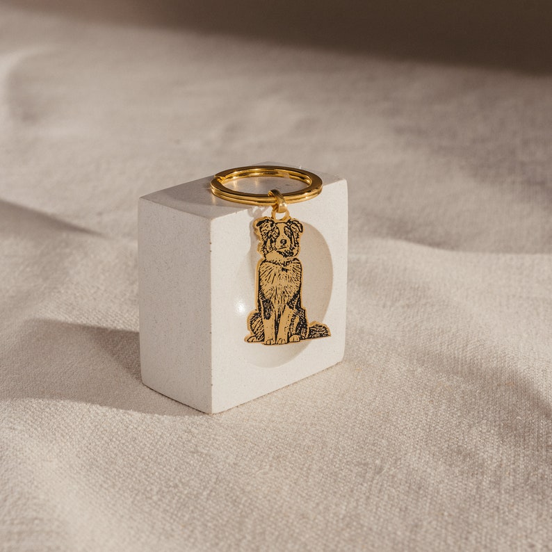 May include: A gold-colored metal keychain with a black and white engraved image of a dog sitting. The keychain is on a white square base.