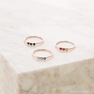 Puede incluir: Tres anillos finos de oro rosa, cada uno con un trío de pequeñas piedras preciosas de corte redondo. Un anillo tiene tres piedras negras, otro piedras roja, verde y amarilla, y el tercero piedras transparente, azul y morada. Texto CAITLYN MINIMALIST.