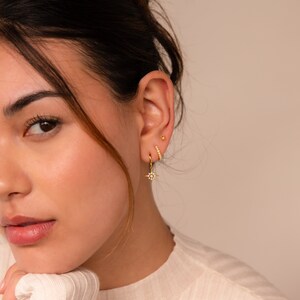 May include: A woman wearing gold earrings with a star design and a small hoop earring. The earrings are set with small white stones.