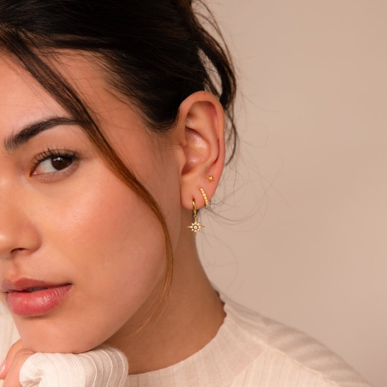 May include: A woman wearing gold earrings with a star design and a small hoop earring. The earrings are set with small white stones.