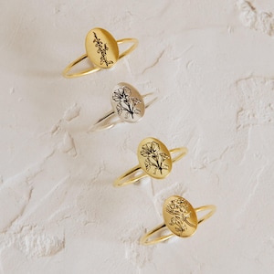 May include: Four rings with oval faces, each featuring a distinct black engraved floral design. Three rings are gold-toned and one is silver-toned. Each ring has a thin, smooth band and a flat, oval-shaped setting displaying a unique botanical illustration.