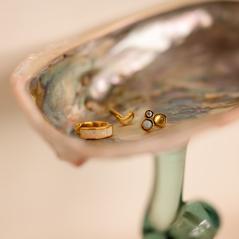 May include: Three gold earrings with opal and diamond accents displayed on a mother of pearl shell. The earrings are a crescent moon, a small bar with an opal, and a cluster of three opals.