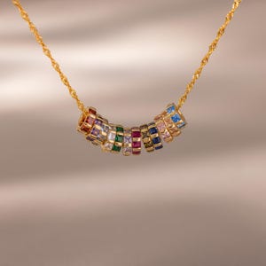 May include: A gold chain necklace featuring a series of small, cylindrical beads, each set with square-cut multicolored gemstones including red, purple, white, green, pink, blue, and yellow stones arranged in a curved, horizontal row.