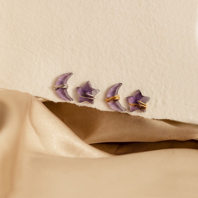 May include: Four amethyst crystal charms, two crescent moons and two stars, wrapped in gold and silver wire, arranged on a beige surface.