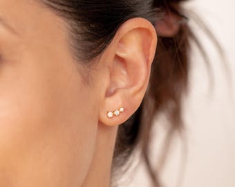 May include: A gold-toned ear climber earring featuring a small, round, iridescent white opal stone followed by two clear, sparkling round-cut stones arranged in a linear, ascending design along the lower edge of the earlobe.
