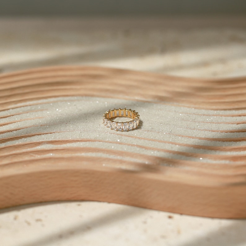 May include: A gold ring with a row of clear baguette-shaped stones set in a continuous band. The ring is resting on a light brown wooden surface.