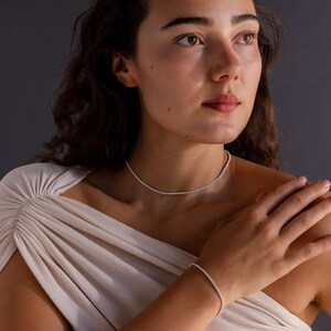 May include: A woman wearing a delicate necklace and bracelet set. The jewelry features a continuous line of small, sparkling stones, creating a refined and elegant look. The pieces are set against a neutral background, highlighting their brilliance.