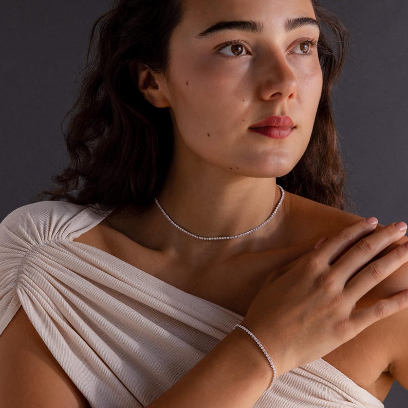 May include: A woman wearing a delicate necklace and bracelet set. The jewelry features a continuous line of small, sparkling stones, creating a refined and elegant look. The pieces are set against a neutral background, highlighting their brilliance.