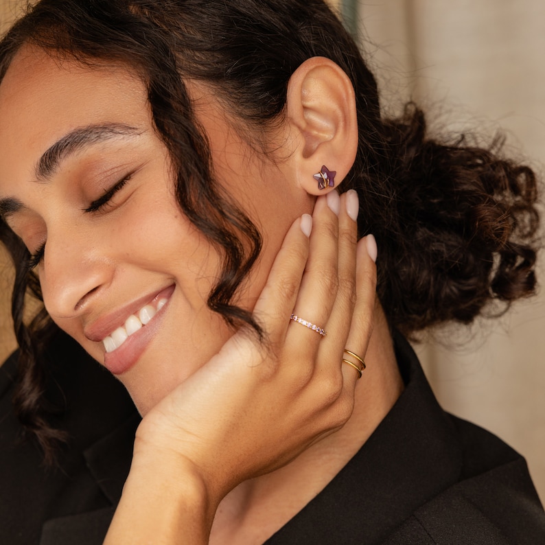May include: A woman wearing a black shirt with a gold ring with pink stones and a gold ring with a simple design. She is also wearing a pair of small star-shaped earrings.