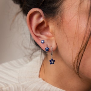 May include: A pair of silver earrings with blue sapphire flower designs. The smaller earring is a stud, while the larger earring is a hoop with a dangling flower charm.