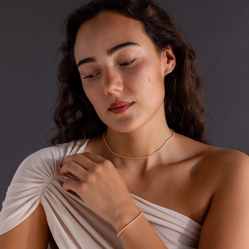 May include: A gold beaded necklace and bracelet set. The jewelry is delicate and appears to be made of small, round beads. The woman in the image is wearing the jewelry. The background is a solid, dark gray.
