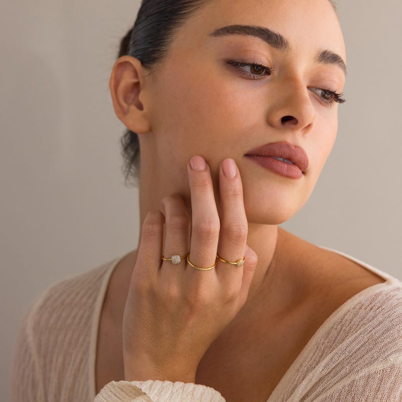 May include: A woman's hand wearing three gold rings. The ring on the index finger has a square-shaped stone with small diamonds. The other two rings are simple bands.