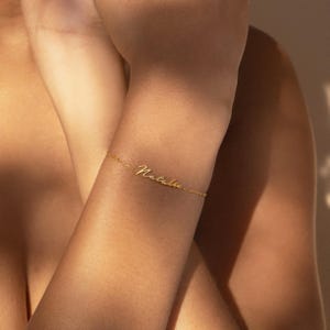 May include: A delicate gold bracelet inscribed with the name "Natalie" in cursive. The bracelet is thin and elegant, with a simple chain design. The bracelet is worn on a wrist.