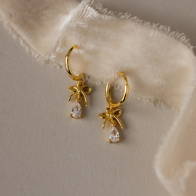 May include: A pair of gold-colored hoop earrings. Each earring features a bow-shaped charm and a teardrop-shaped, clear gemstone pendant. The earrings are displayed on a neutral background with a soft, white fabric.