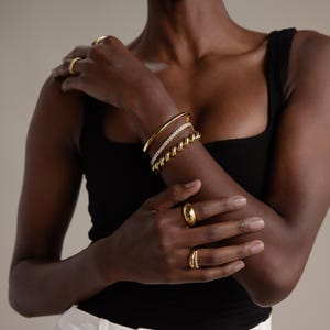 May include: A selection of gold jewellery, including rings and bracelets, is shown on a person's arm and fingers. The bracelets include a solid gold bangle, a diamond-set band, and a twisted rope design. The rings are various styles, including a dome ring.