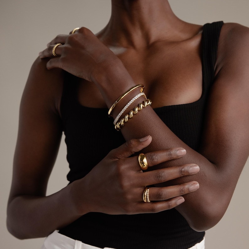 May include: A selection of gold jewellery, including rings and bracelets, is shown on a person's arm and fingers. The bracelets include a solid gold bangle, a diamond-set band, and a twisted rope design. The rings are various styles, including a dome ring.