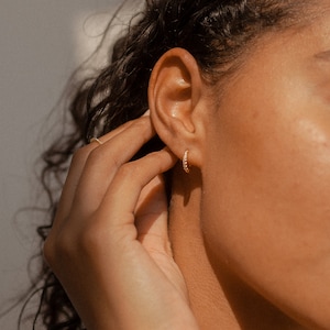 May include: Close-up of a gold hoop earring with small, clear stones. The earring is being held near an ear. The skin tone is warm, and the lighting is soft. The earring is a classic jewelry piece.