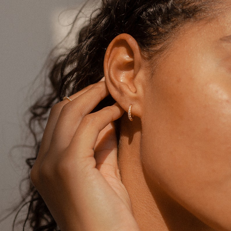 May include: Close-up of a gold hoop earring with small, clear stones. The earring is being held near an ear. The skin tone is warm, and the lighting is soft. The earring is a classic jewelry piece.