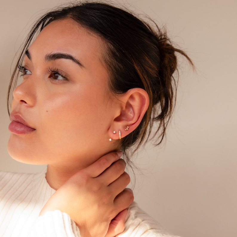 May include: A close-up of a person's ear adorned with three sets of gold earrings. The earrings include small studs and a bar-shaped earring with multiple small stones. The skin tone is light, and the background is a soft, neutral color.