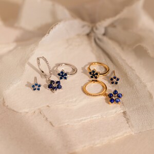 May include: A set of silver and gold earrings featuring blue sapphire flower designs. The earrings are displayed on a white textured surface.