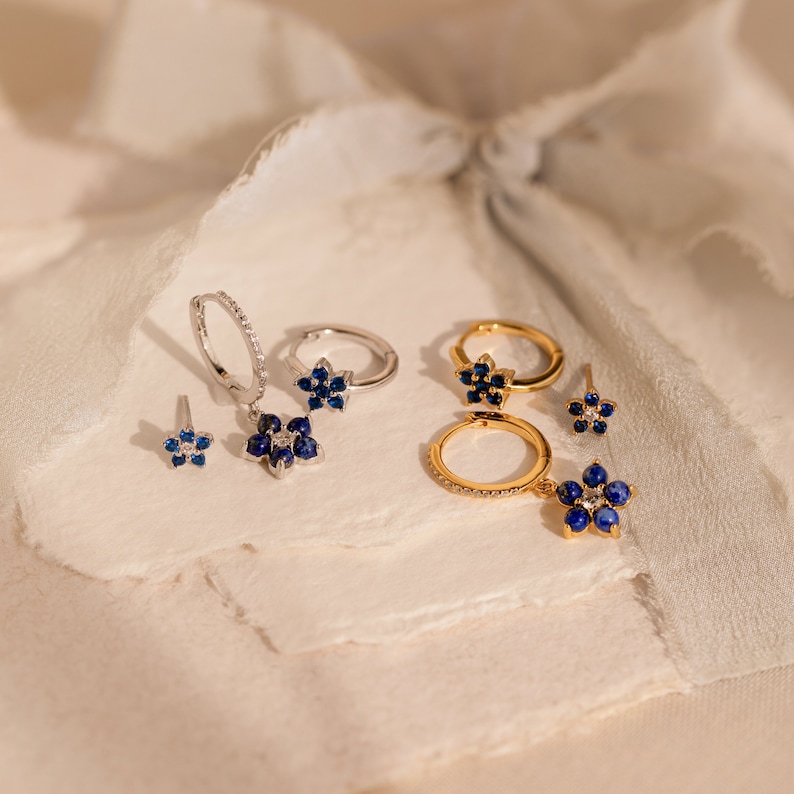 May include: A set of silver and gold earrings featuring blue sapphire flower designs. The earrings are displayed on a white textured surface.