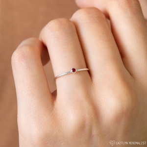 Peut inclure: Une fine bague en argent dotée d'une petite pierre précieuse ronde d'un rouge profond, sertie dans une monture en métal simple. La bague présente une finition lisse et polie ainsi qu'un profil délicat et fin qui entoure le doigt avec élégance.