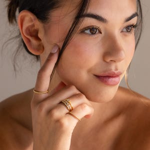 May include: A close-up of a hand wearing several gold rings. The rings include a beaded band, a multi-layered ring with small stones, and a delicate band with tiny stones. The rings are set against a soft, neutral background.