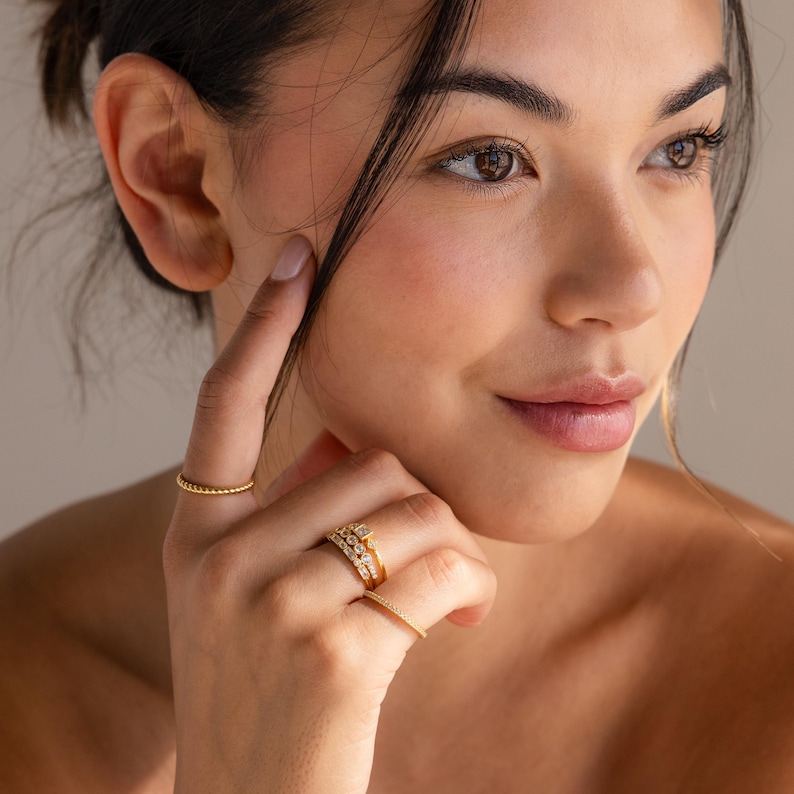 May include: A close-up of a hand wearing several gold rings. The rings include a beaded band, a multi-layered ring with small stones, and a delicate band with tiny stones. The rings are set against a soft, neutral background.
