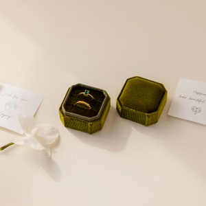 May include: A green velvet ring box with two gold rings and a green gemstone ring inside. The box is open and the rings are visible. There is a card with the text "Happiness looks beautiful" on the right side of the image.