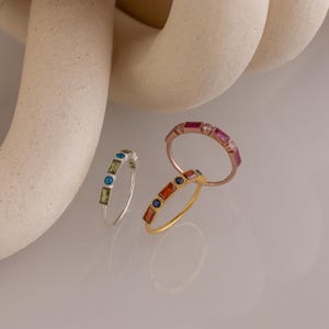 May include: Three gemstone rings in silver, gold, and rose gold. The silver ring has green and blue stones. The gold ring has orange and blue stones. The rose gold ring has pink and white stones. The rings are displayed on a reflective surface.